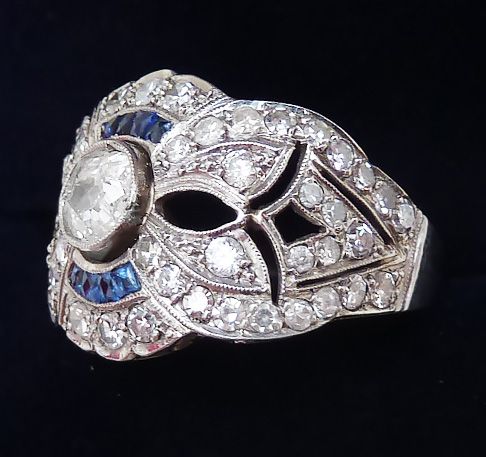 fig 2. An Art Deco platinum cluster ring featuring a central round diamond surrounded by blue sapphire arcs, geometric openwork, and smaller accent diamonds.