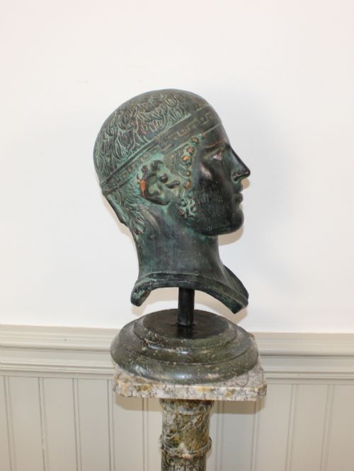 fig 1. A dark green patinated metal portrait bust depicting a classical male head with a patterned headband resting on a circular stepped base.