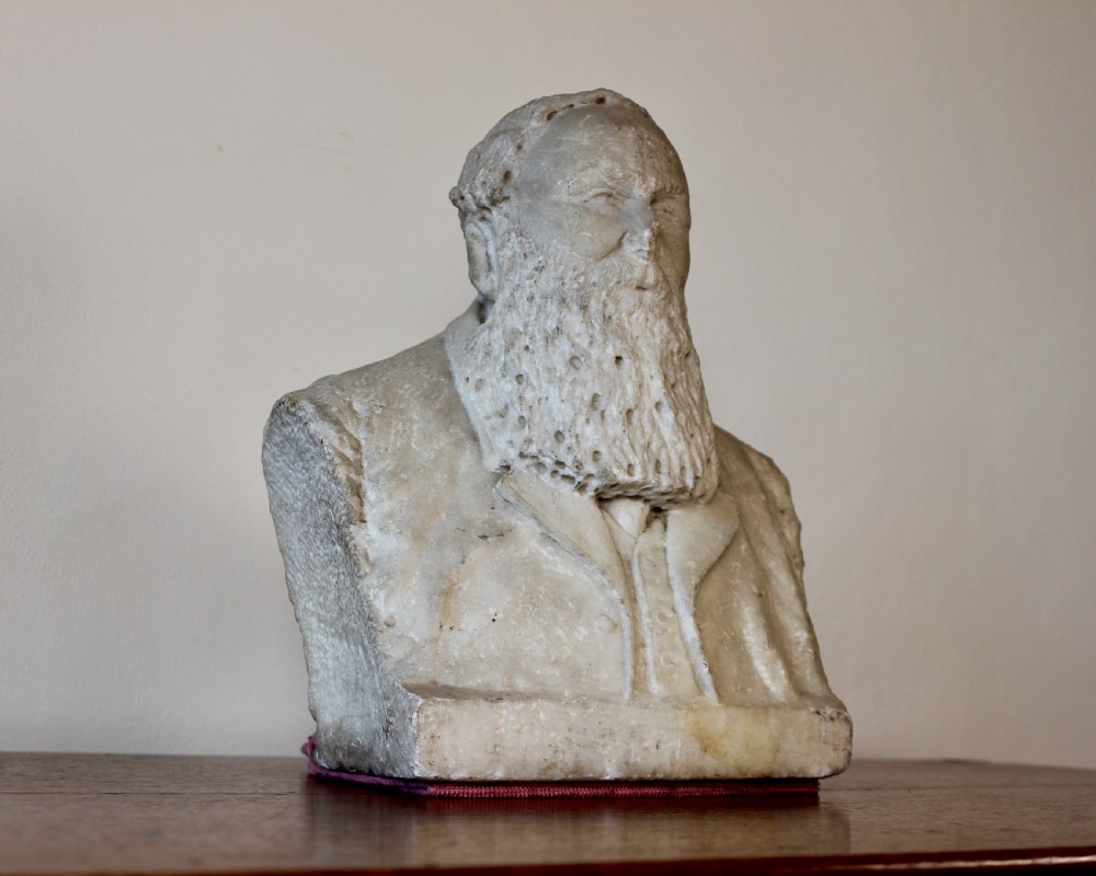 A carved off-white marble bust representing a bearded man wearing a Victorian jacket and waistcoat, set on an integrated square plinth base.
