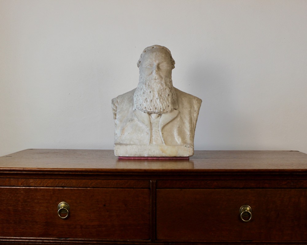 A carved off-white marble bust representing a bearded man wearing a Victorian jacket and waistcoat, set on an integrated square plinth base. View II