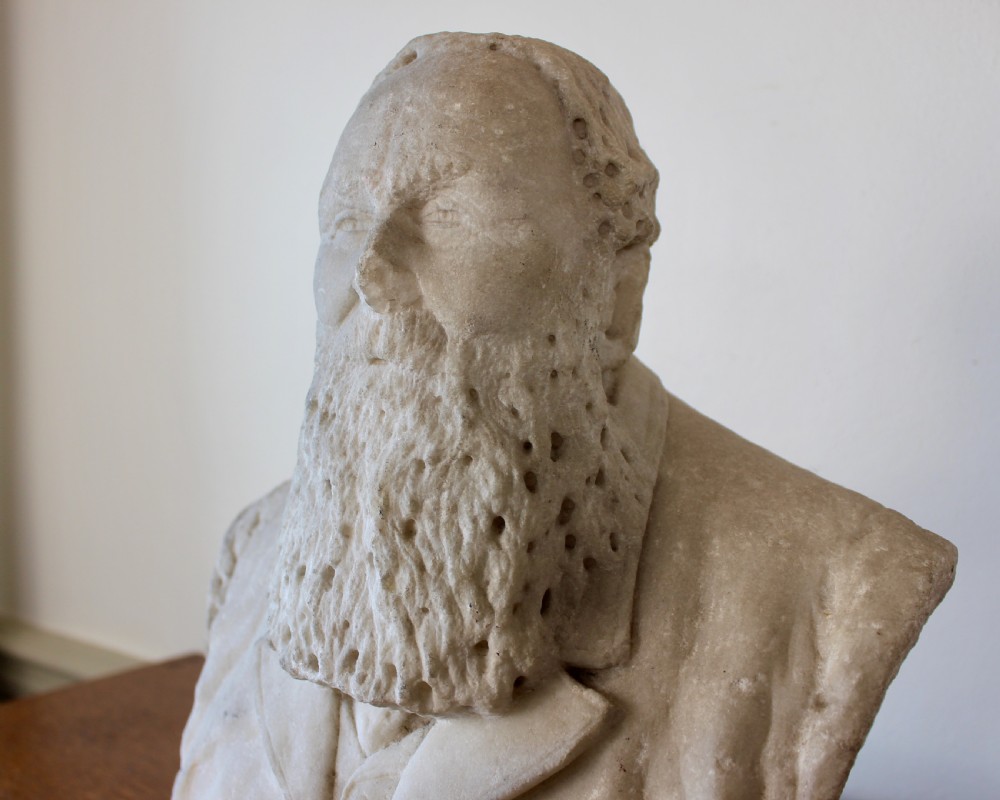 A carved off-white marble bust representing a bearded man wearing a Victorian jacket and waistcoat, set on an integrated square plinth base. View IV