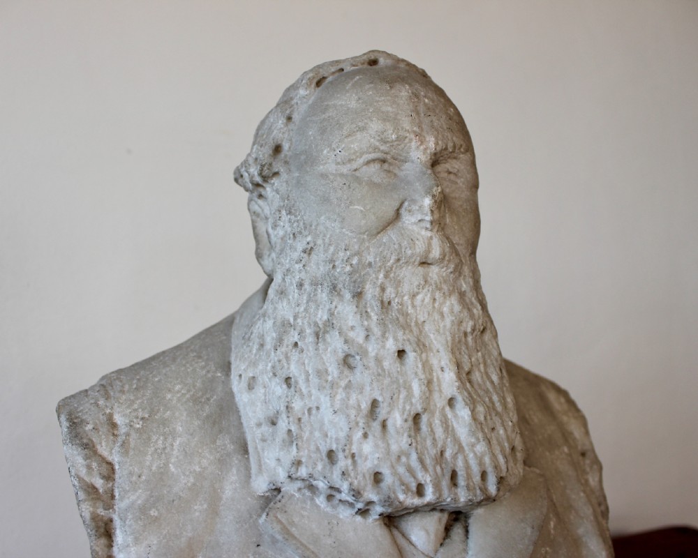 A carved off-white marble bust representing a bearded man wearing a Victorian jacket and waistcoat, set on an integrated square plinth base. View V