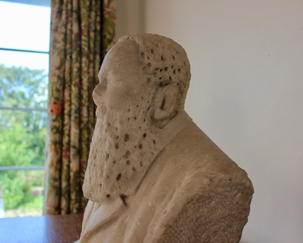 A carved off-white marble bust representing a bearded man wearing a Victorian jacket and waistcoat, set on an integrated square plinth base. View VI