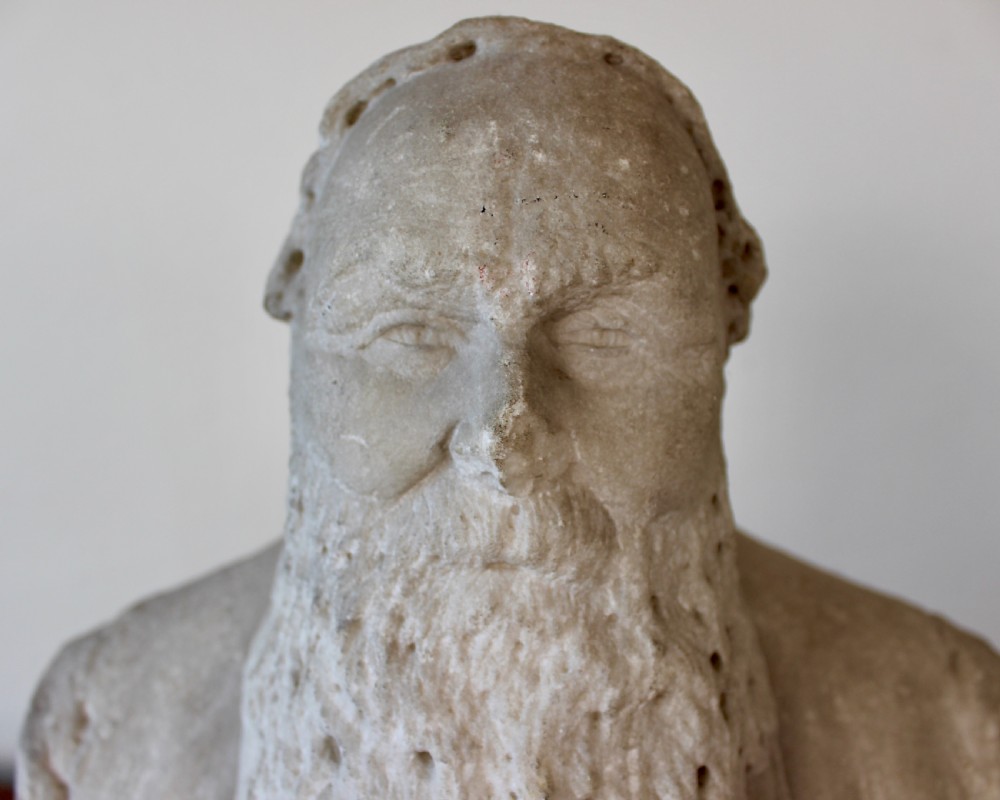 A carved off-white marble bust representing a bearded man wearing a Victorian jacket and waistcoat, set on an integrated square plinth base. View VII