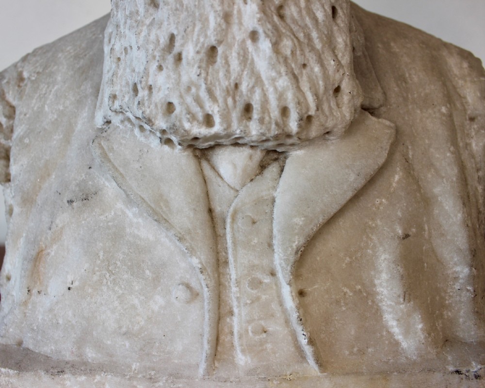 A carved off-white marble bust representing a bearded man wearing a Victorian jacket and waistcoat, set on an integrated square plinth base. View VIII