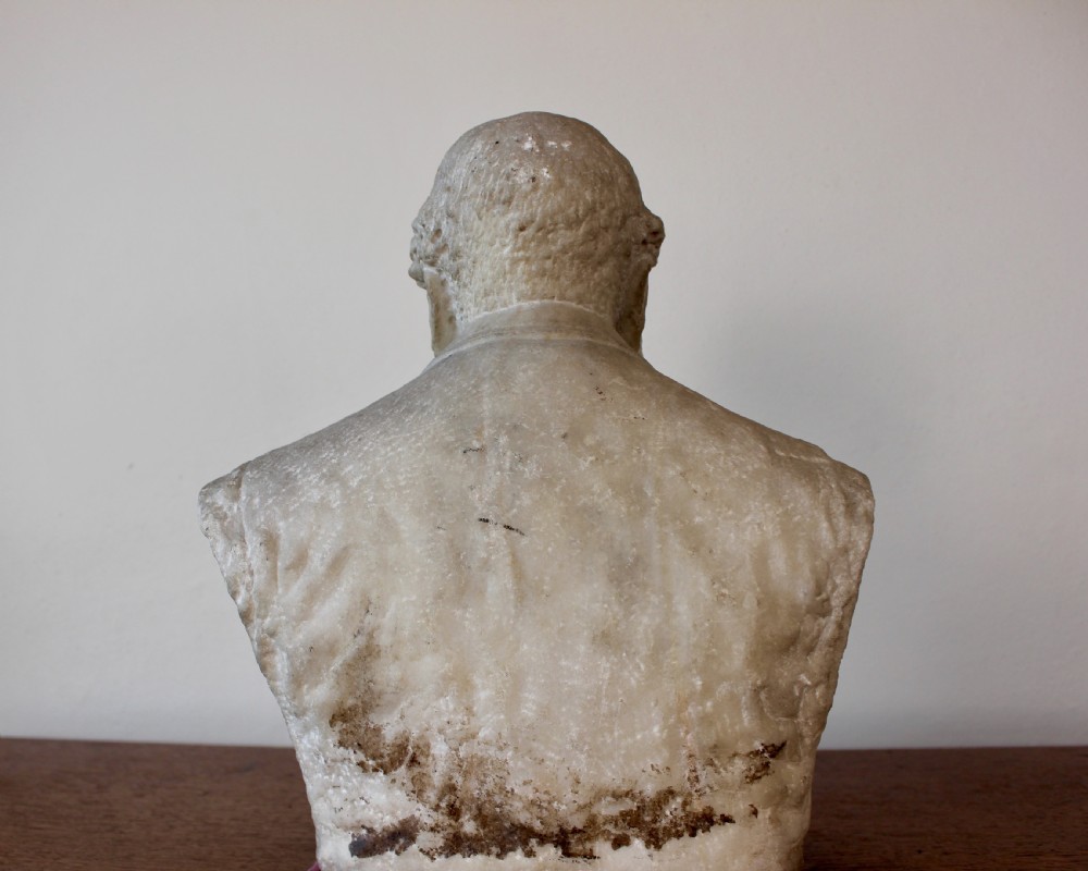 A carved off-white marble bust representing a bearded man wearing a Victorian jacket and waistcoat, set on an integrated square plinth base. View IX