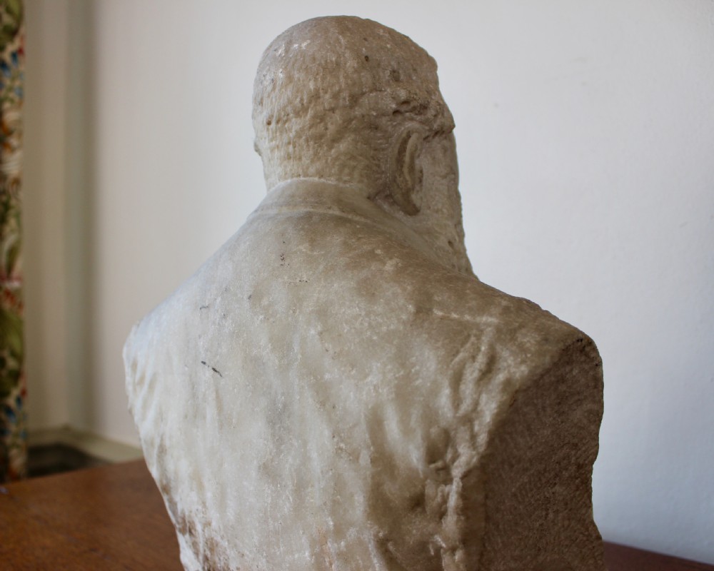 A carved off-white marble bust representing a bearded man wearing a Victorian jacket and waistcoat, set on an integrated square plinth base. View X