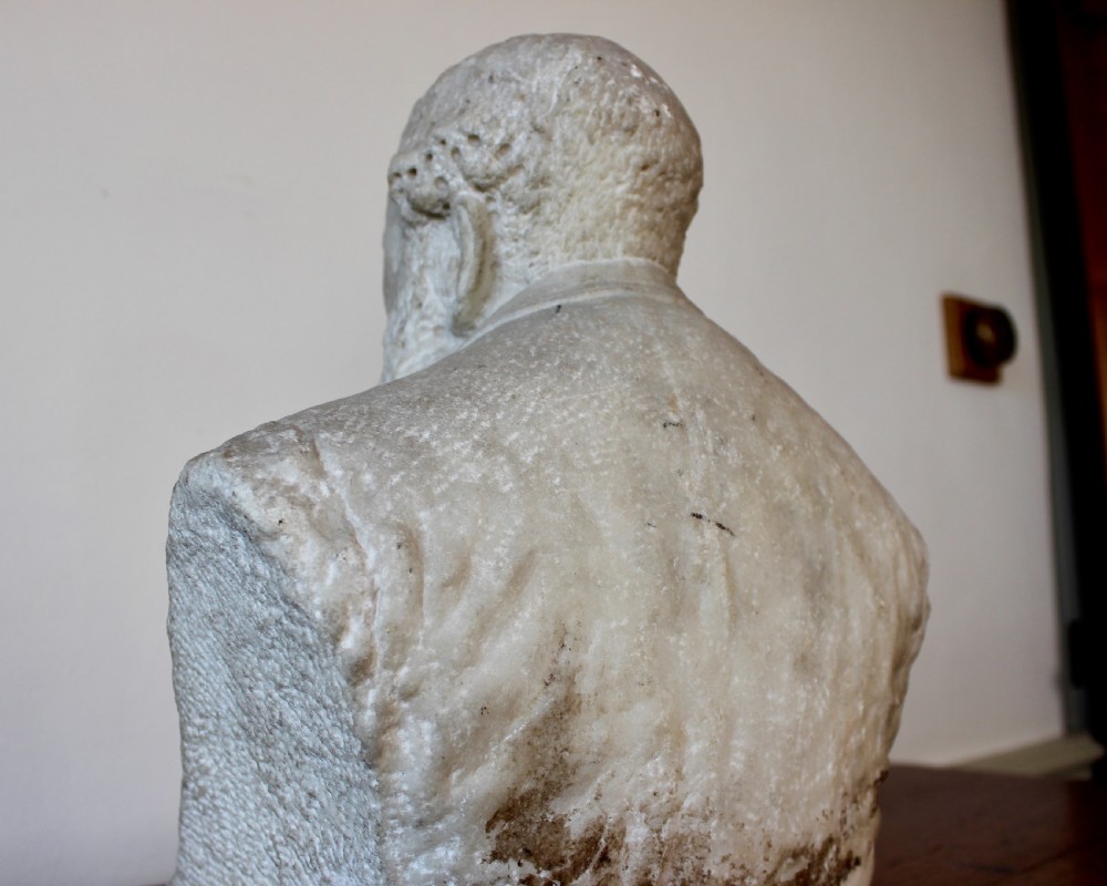 A carved off-white marble bust representing a bearded man wearing a Victorian jacket and waistcoat, set on an integrated square plinth base. View XI