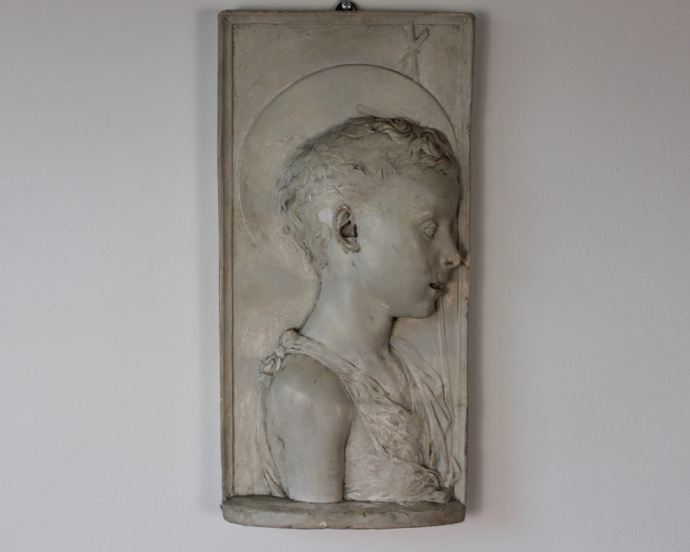 A rectangular plaster bas-relief wall plaque depicting the profile portrait of a young saint with a halo and cross motif.