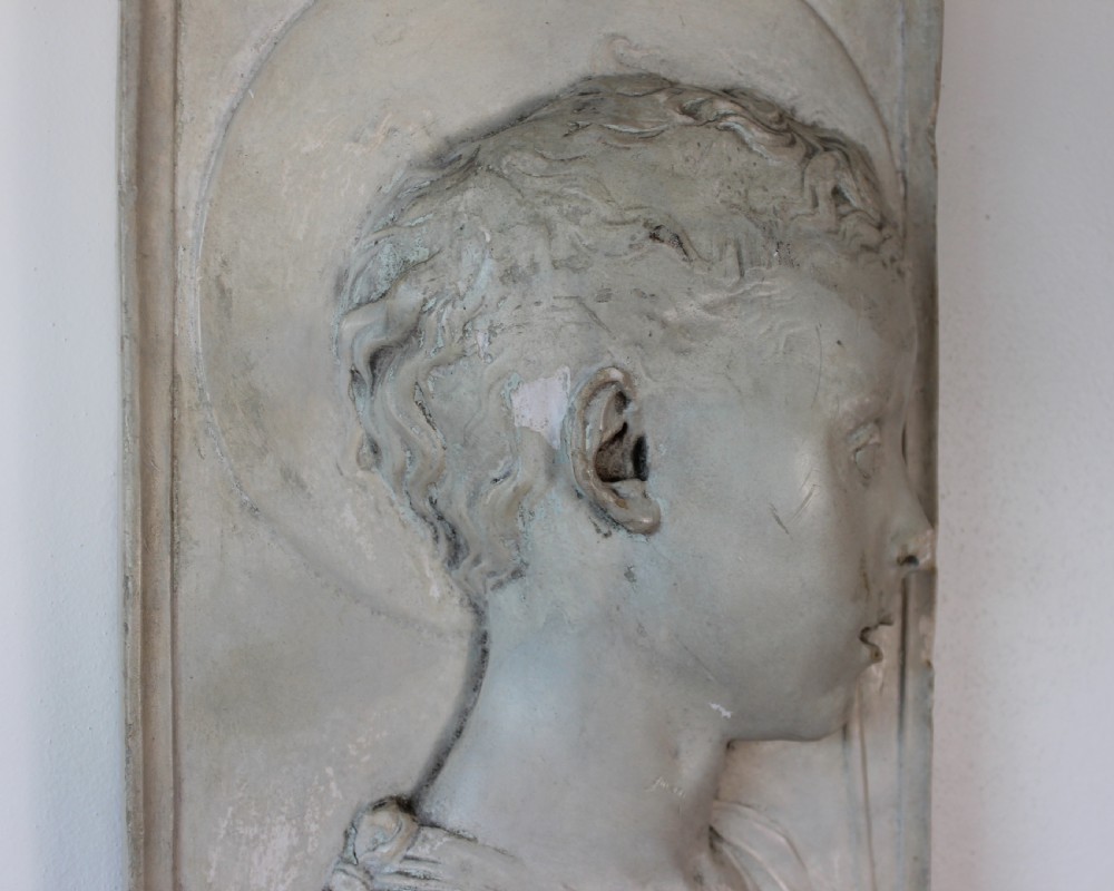 A rectangular plaster bas-relief wall plaque depicting the profile portrait of a young saint with a halo and cross motif. View II