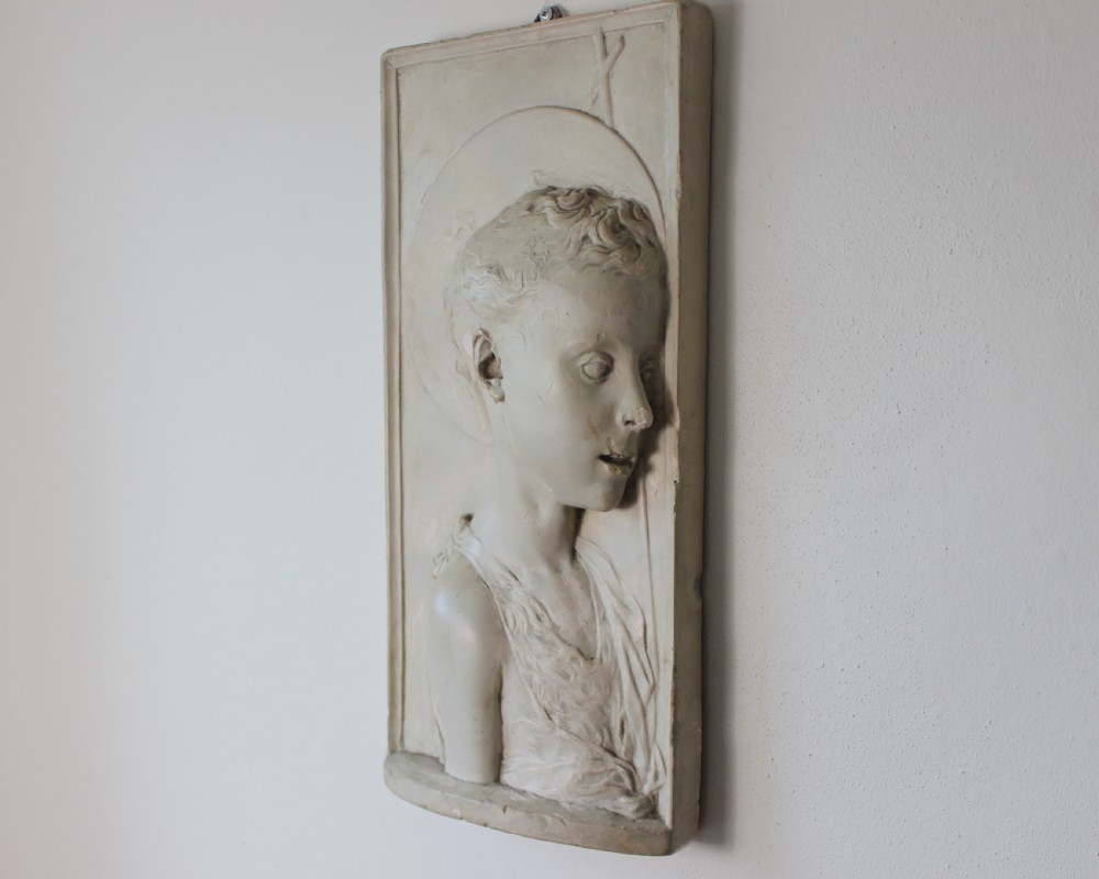 A rectangular plaster bas-relief wall plaque depicting the profile portrait of a young saint with a halo and cross motif. View III