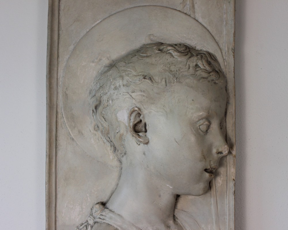 A rectangular plaster bas-relief wall plaque depicting the profile portrait of a young saint with a halo and cross motif. View IV