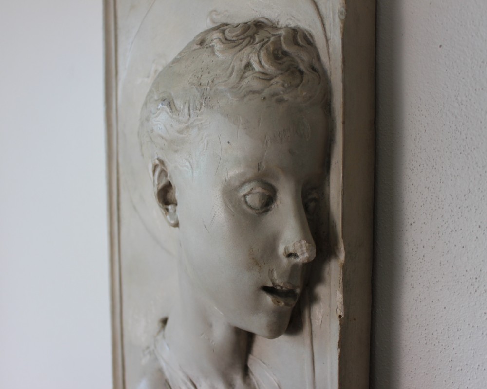 A rectangular plaster bas-relief wall plaque depicting the profile portrait of a young saint with a halo and cross motif. View V
