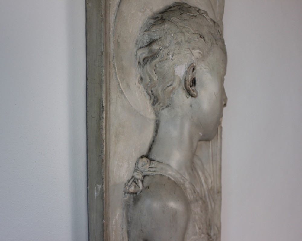 A rectangular plaster bas-relief wall plaque depicting the profile portrait of a young saint with a halo and cross motif. View VI