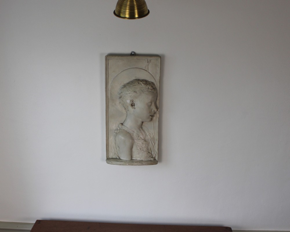 A rectangular plaster bas-relief wall plaque depicting the profile portrait of a young saint with a halo and cross motif. View VII
