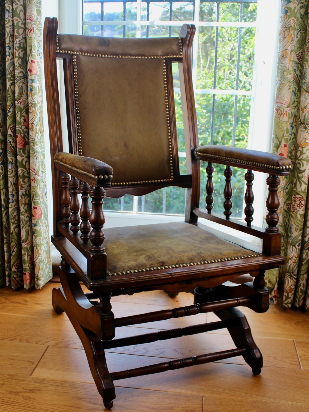 An Edwardian mahogany platform rocking chair with patinated leather upholstery, brass stud detail, and turned spindles. View IV