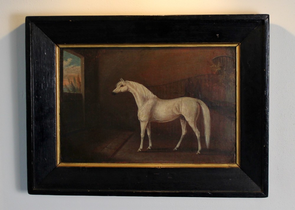 An early 19th-century oil painting depicting a white stallion in profile inside a dimly lit stable, presented in a black ebonised frame with a gilded inner slip.