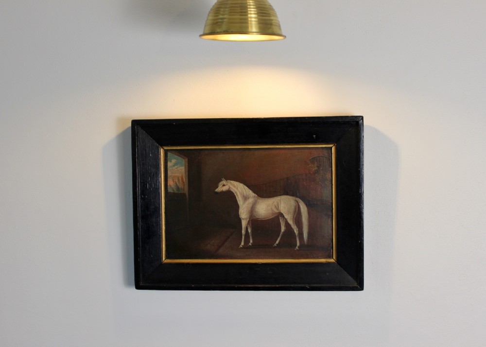 An early 19th-century oil painting depicting a white stallion in profile inside a dimly lit stable, presented in a black ebonised frame with a gilded inner slip. View V