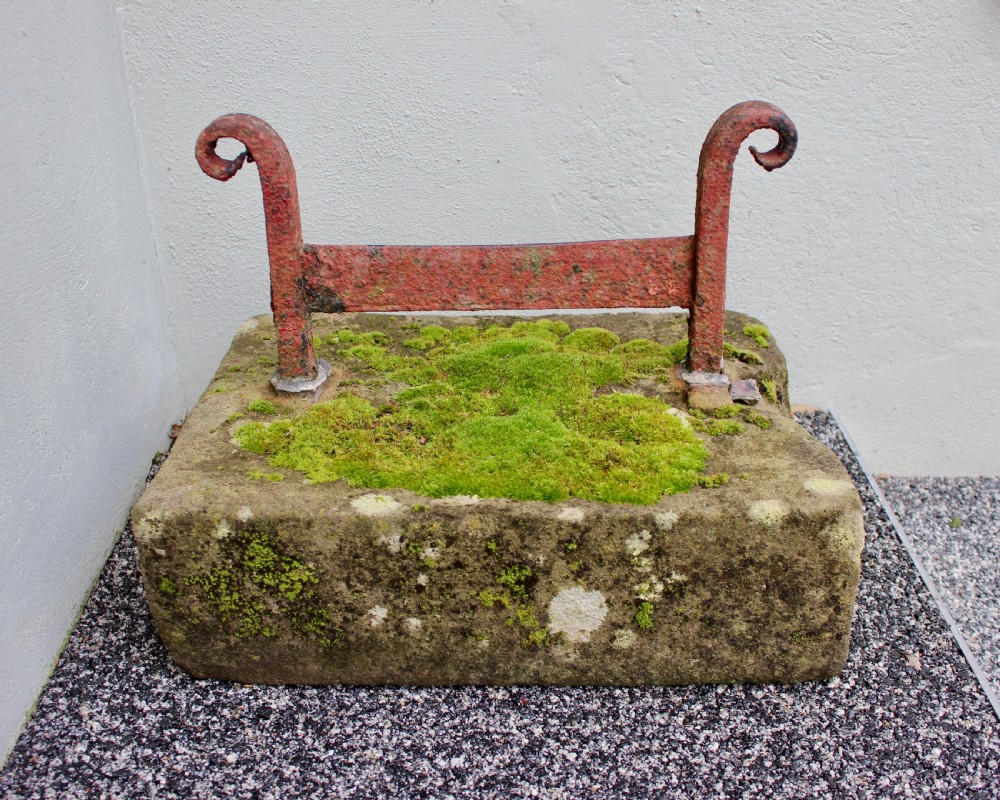 A late Georgian boot scraper featuring a wrought iron blade with scrolled side supports, anchored into a rectangular weathered stone block covered in moss.