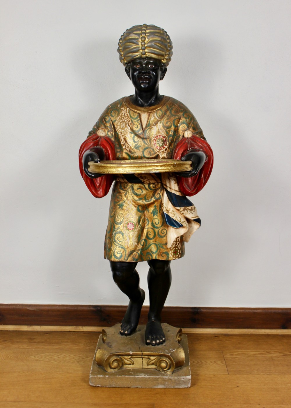 A carved wooden standing Venetian waiter figure wearing a decorated gold tunic and turban while holding a tray on a carved base.