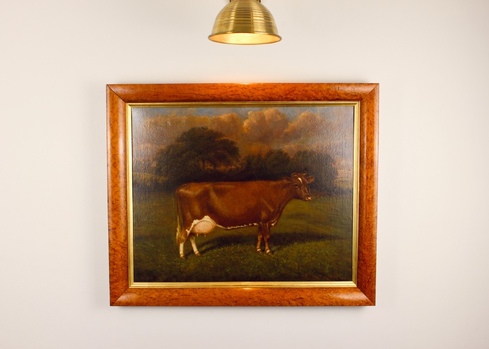 An Edwardian oil painting depicts a brown cow standing in a grassy field before trees, presented within a wide figured maple wood frame with a gold slip. View II