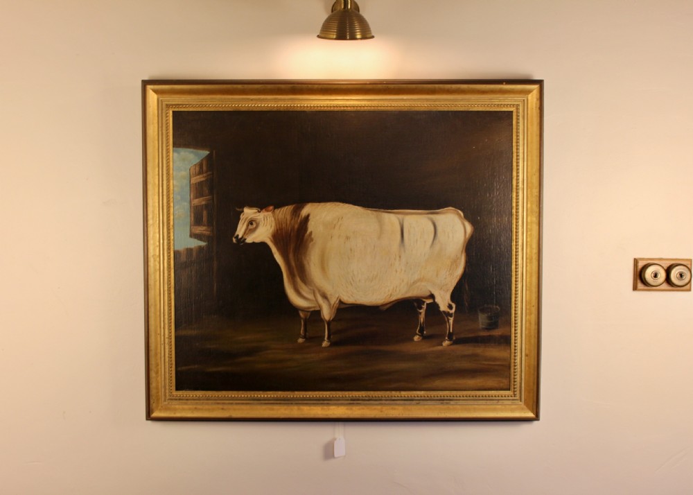 A 19th-century naive oil painting depicting a white and brown cow standing inside a dark stable framed in a gilded timber frame.