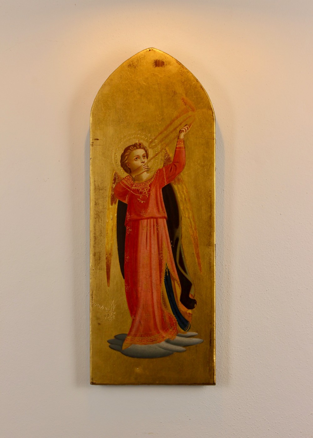 A tall arched wooden panel featuring gold leaf gilding and a central painted figure of an angel wearing a red robe and holding a long trumpet.