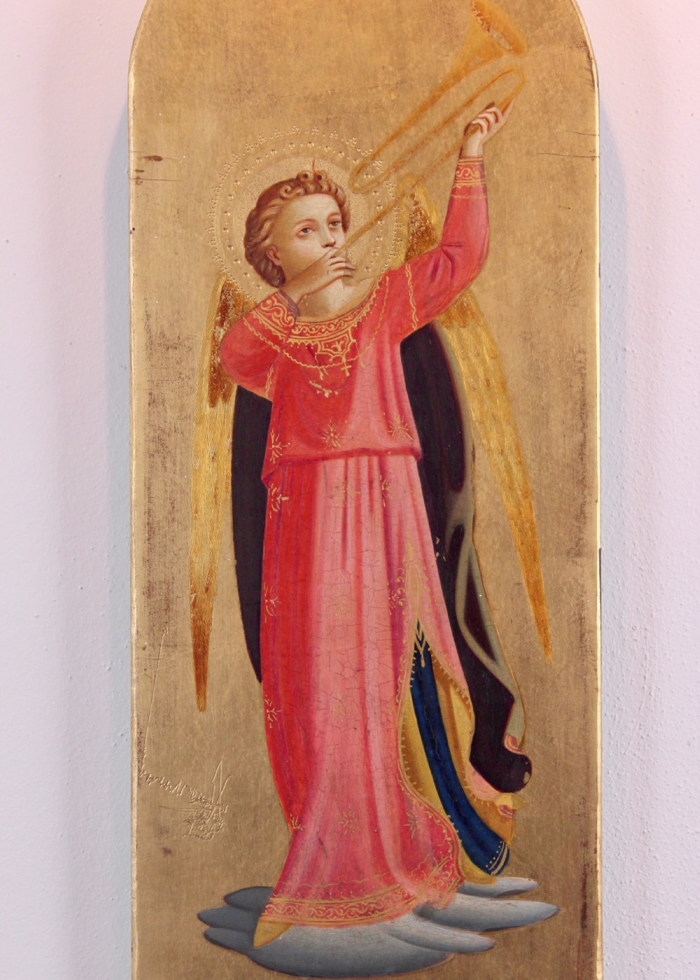 A tall arched wooden panel featuring gold leaf gilding and a central painted figure of an angel wearing a red robe and holding a long trumpet. View III