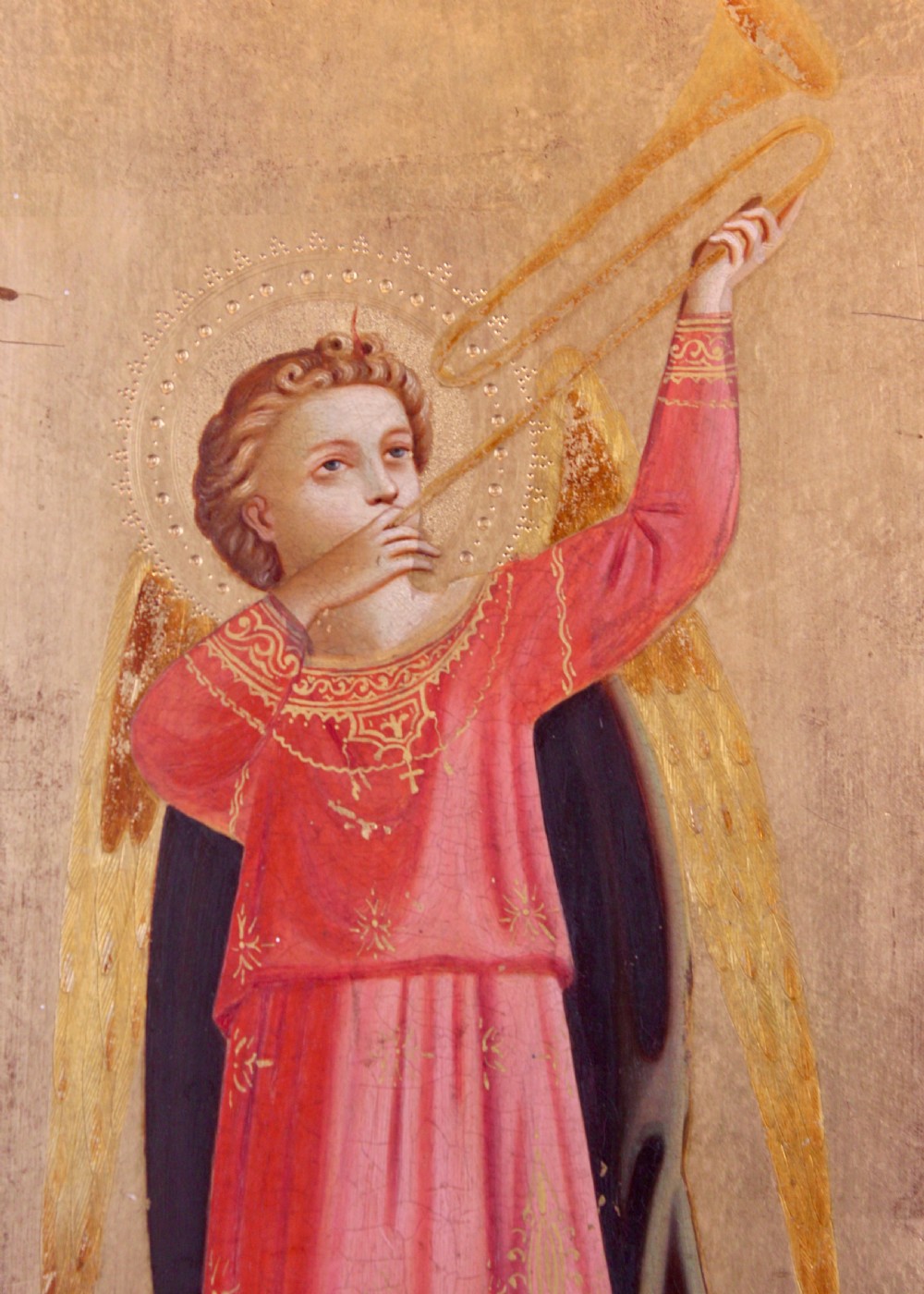 A tall arched wooden panel featuring gold leaf gilding and a central painted figure of an angel wearing a red robe and holding a long trumpet. View V