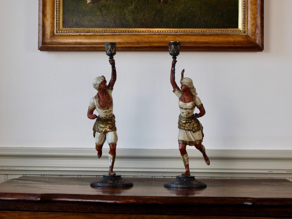 A pair of antique cold-painted spelter figural candlesticks depicting male and female figures in Orientalist attire holding raised torch nozzles.