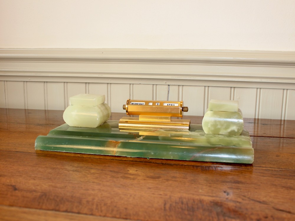 An Art Deco green onyx desk stand featuring a central gilt brass perpetual calendar flanked by two lidded onyx inkwells on a stepped rectangular base with a pen groove.