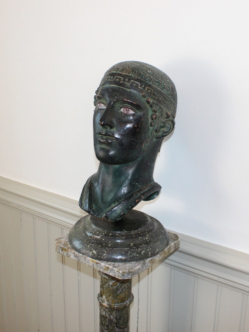 A dark green patinated metal portrait bust depicting a classical male head with a patterned headband resting on a circular stepped base.