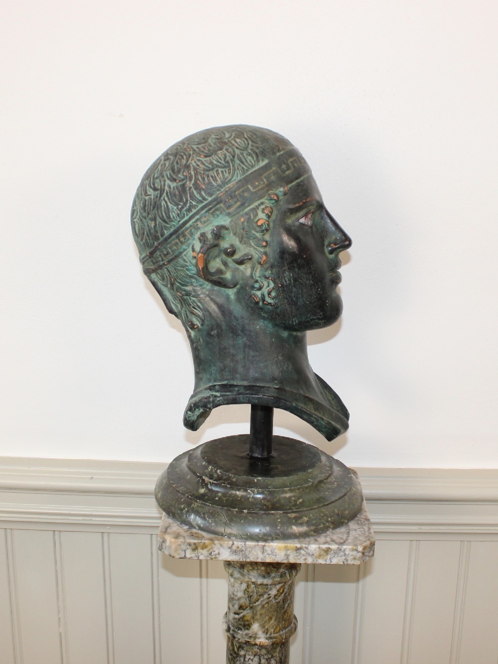 A dark green patinated metal portrait bust depicting a classical male head with a patterned headband resting on a circular stepped base. View II