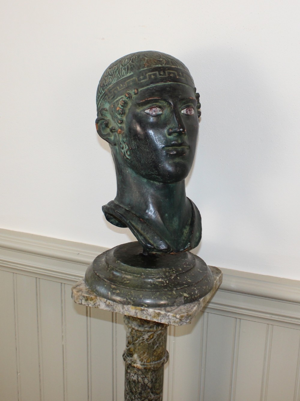 A dark green patinated metal portrait bust depicting a classical male head with a patterned headband resting on a circular stepped base. View III