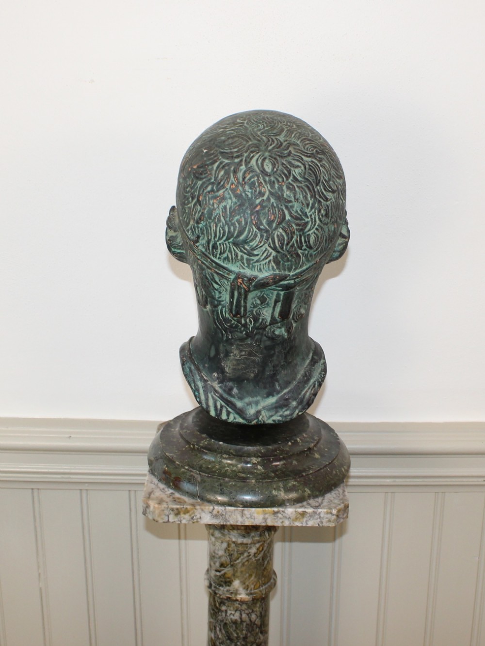 A dark green patinated metal portrait bust depicting a classical male head with a patterned headband resting on a circular stepped base. View IV