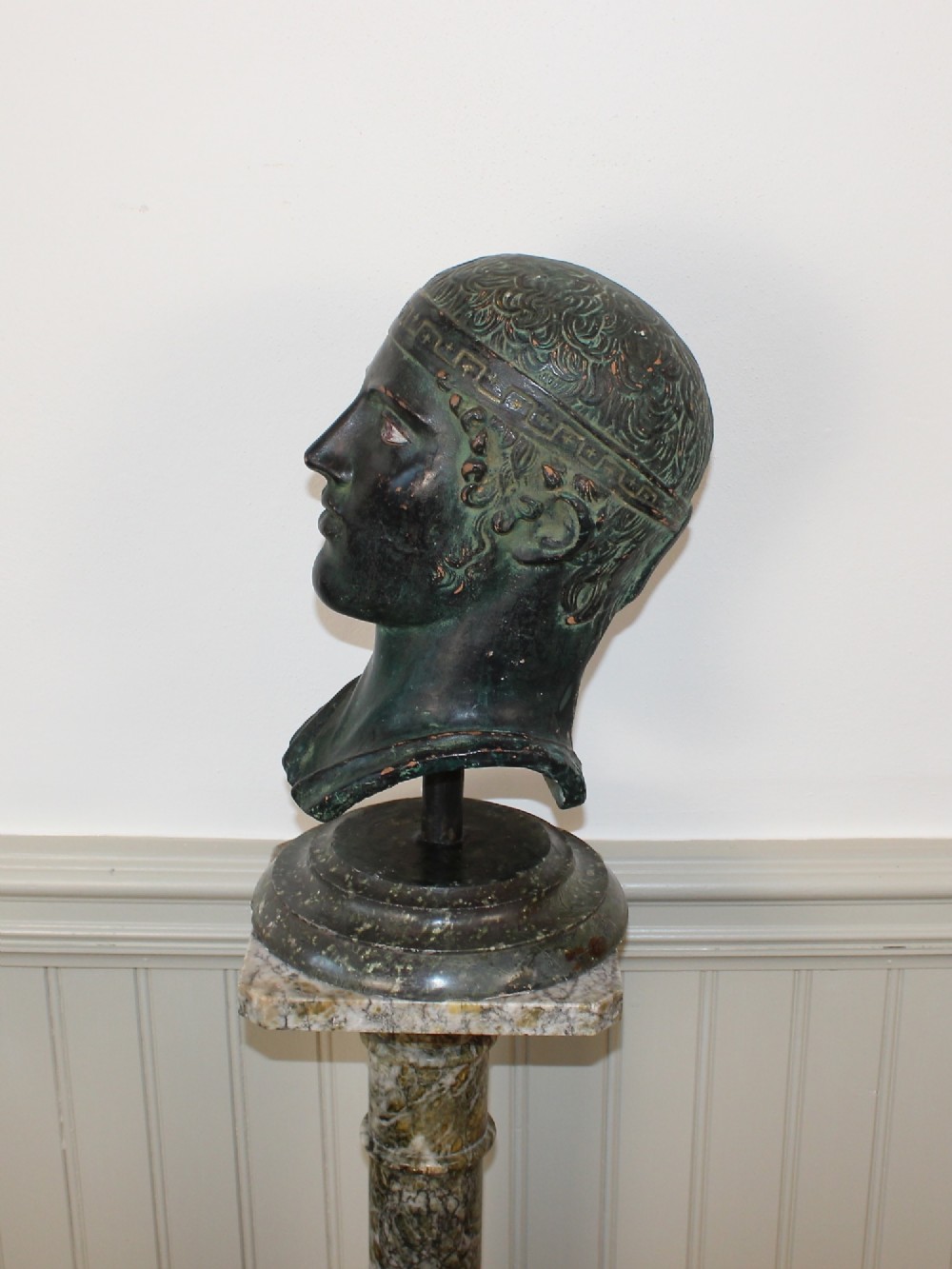 A dark green patinated metal portrait bust depicting a classical male head with a patterned headband resting on a circular stepped base. View V