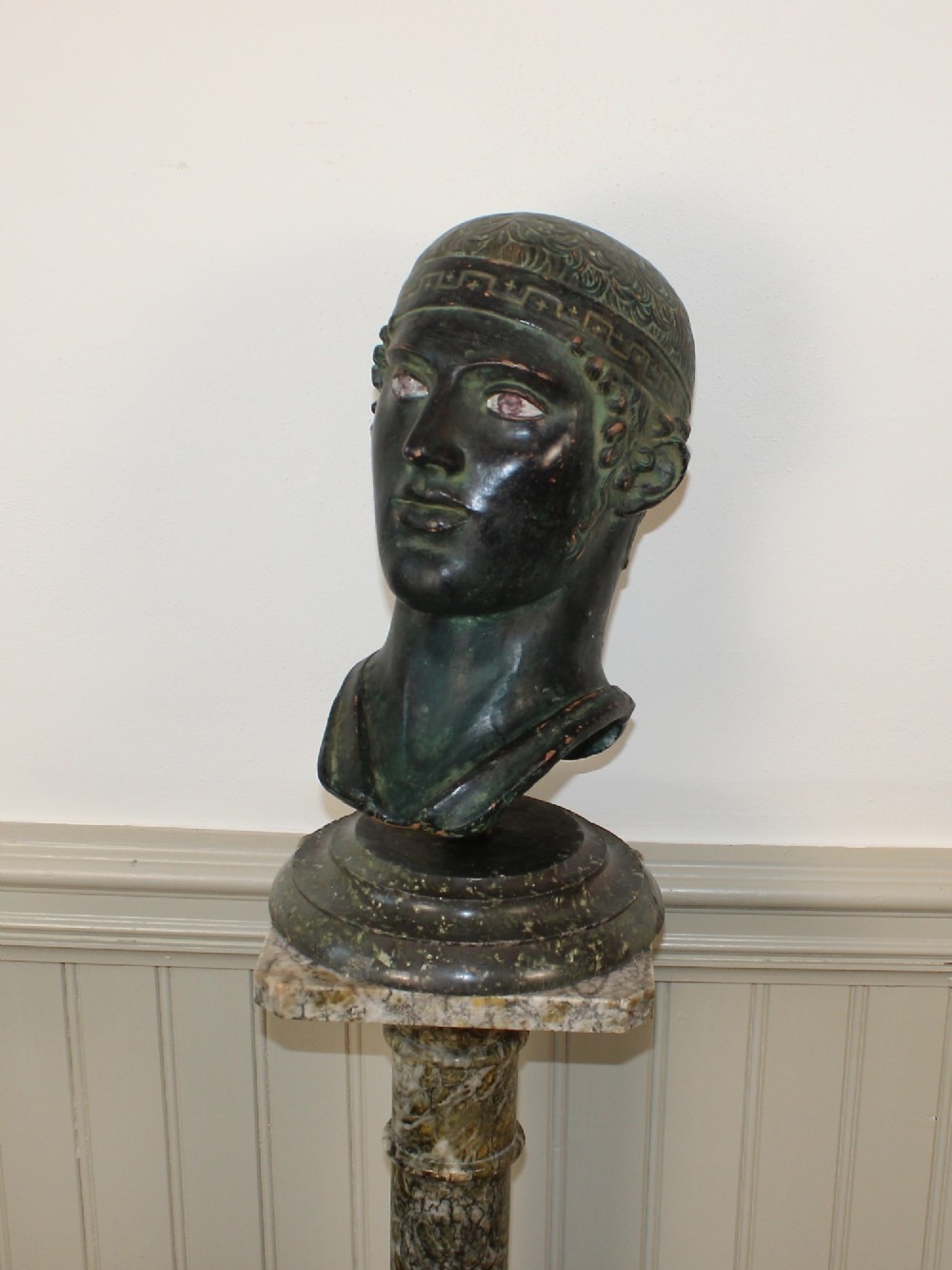 A dark green patinated metal portrait bust depicting a classical male head with a patterned headband resting on a circular stepped base. View VI