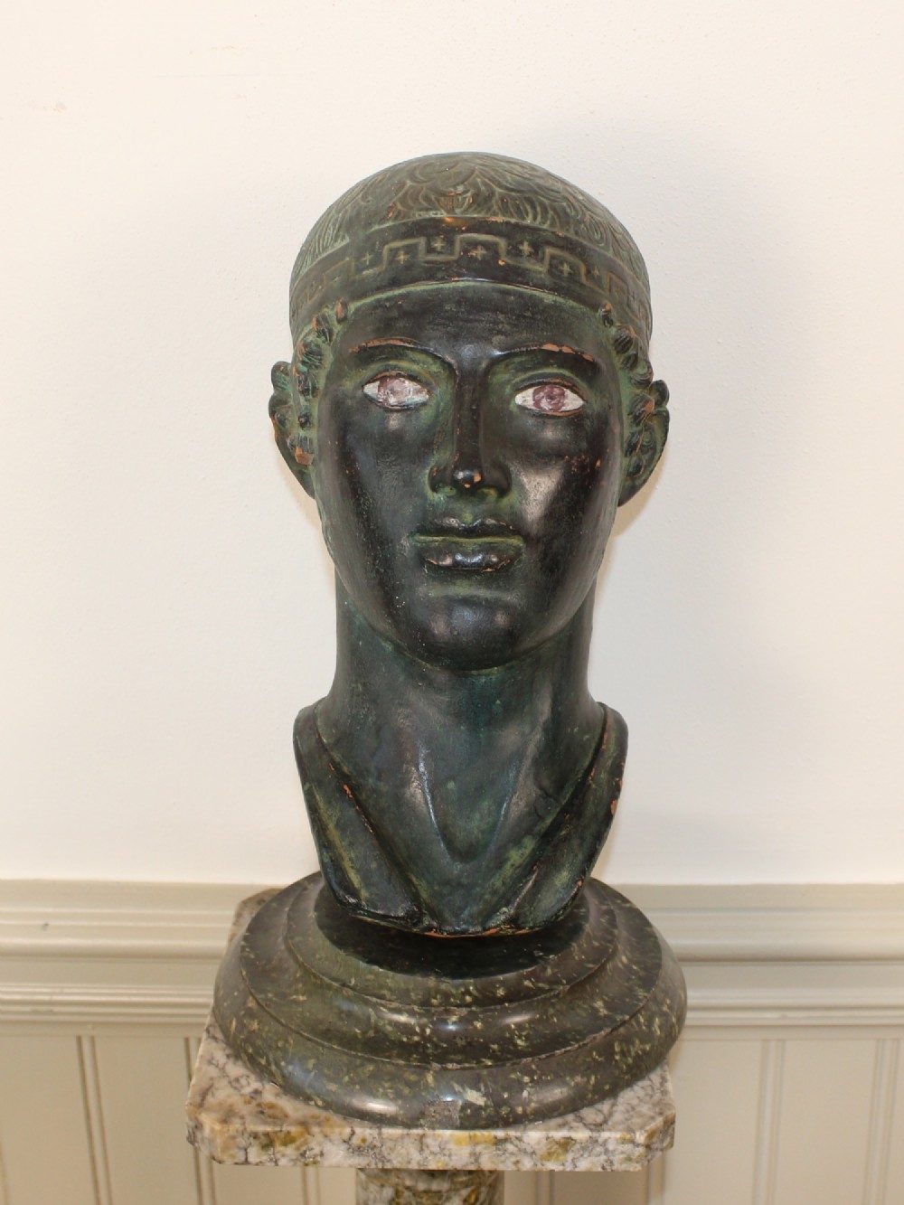 A dark green patinated metal portrait bust depicting a classical male head with a patterned headband resting on a circular stepped base. View VII