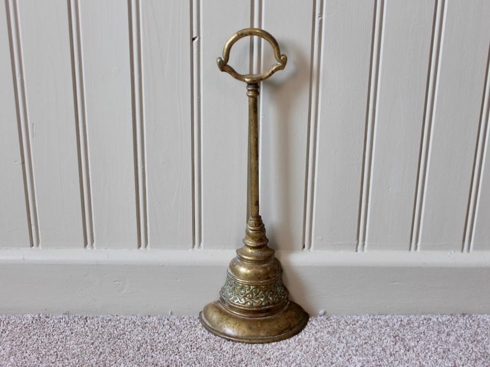 An antique Victorian cast brass door porter featuring a top loop handle, vertical stem, and weighted stepped base decorated with relief scrollwork.
