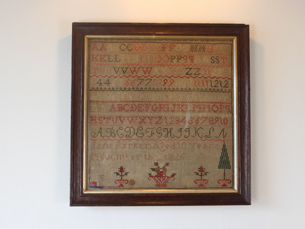 A framed 1825 Regency needlework alphabet sampler on linen fabric featuring embroidered rows of letters, numbers, floral motifs, and an inscription inside a dark timber frame.