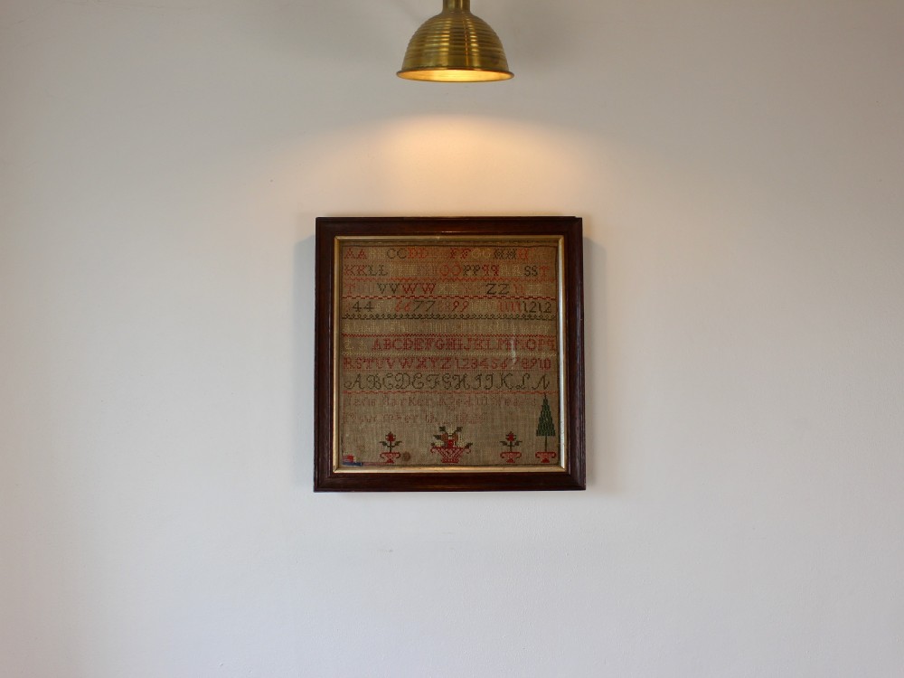 A framed 1825 Regency needlework alphabet sampler on linen fabric featuring embroidered rows of letters, numbers, floral motifs, and an inscription inside a dark timber frame. View II