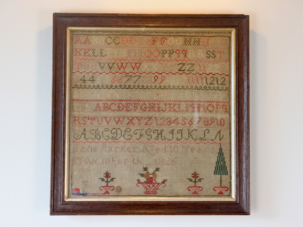 A framed 1825 Regency needlework alphabet sampler on linen fabric featuring embroidered rows of letters, numbers, floral motifs, and an inscription inside a dark timber frame. View III