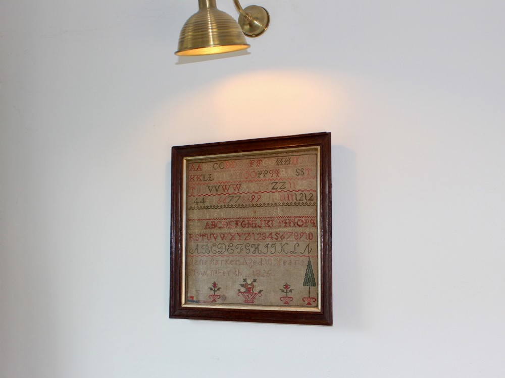 A framed 1825 Regency needlework alphabet sampler on linen fabric featuring embroidered rows of letters, numbers, floral motifs, and an inscription inside a dark timber frame. View IV