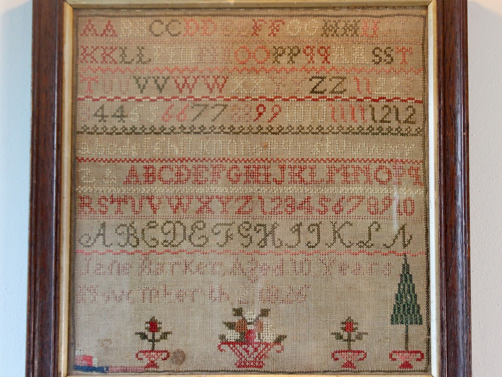 A framed 1825 Regency needlework alphabet sampler on linen fabric featuring embroidered rows of letters, numbers, floral motifs, and an inscription inside a dark timber frame. View V