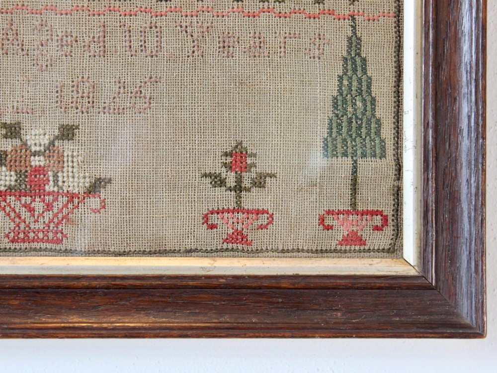 A framed 1825 Regency needlework alphabet sampler on linen fabric featuring embroidered rows of letters, numbers, floral motifs, and an inscription inside a dark timber frame. View VI