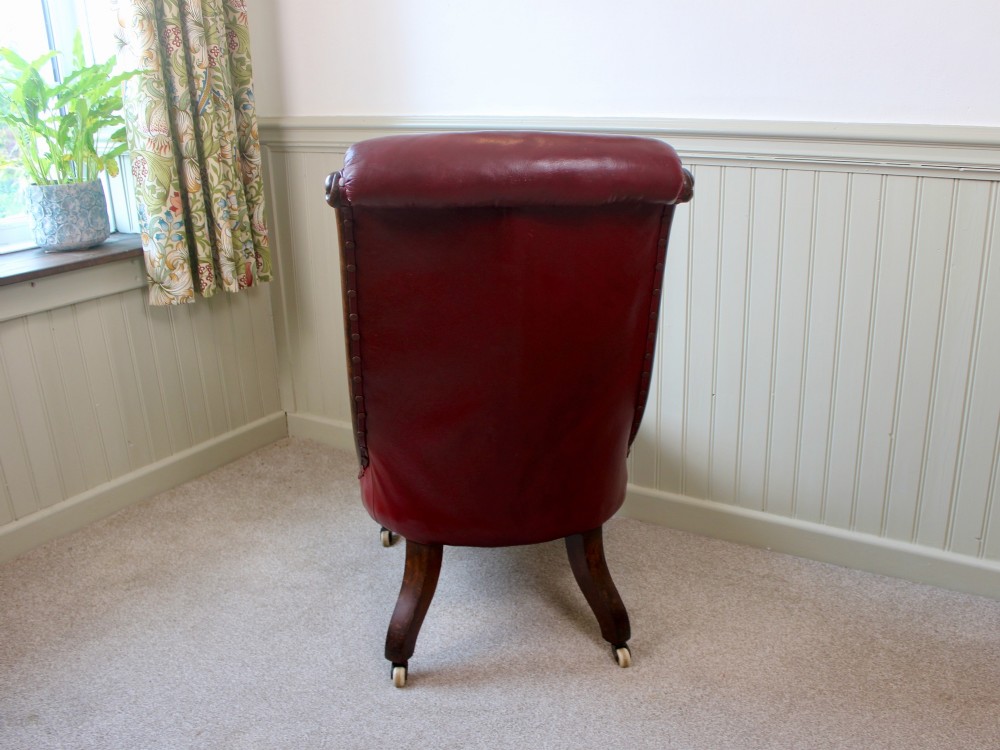 An early Victorian slipper chair featuring deep-buttoned red leather upholstery, a rosewood frame, and turned front legs on brass castors. View V