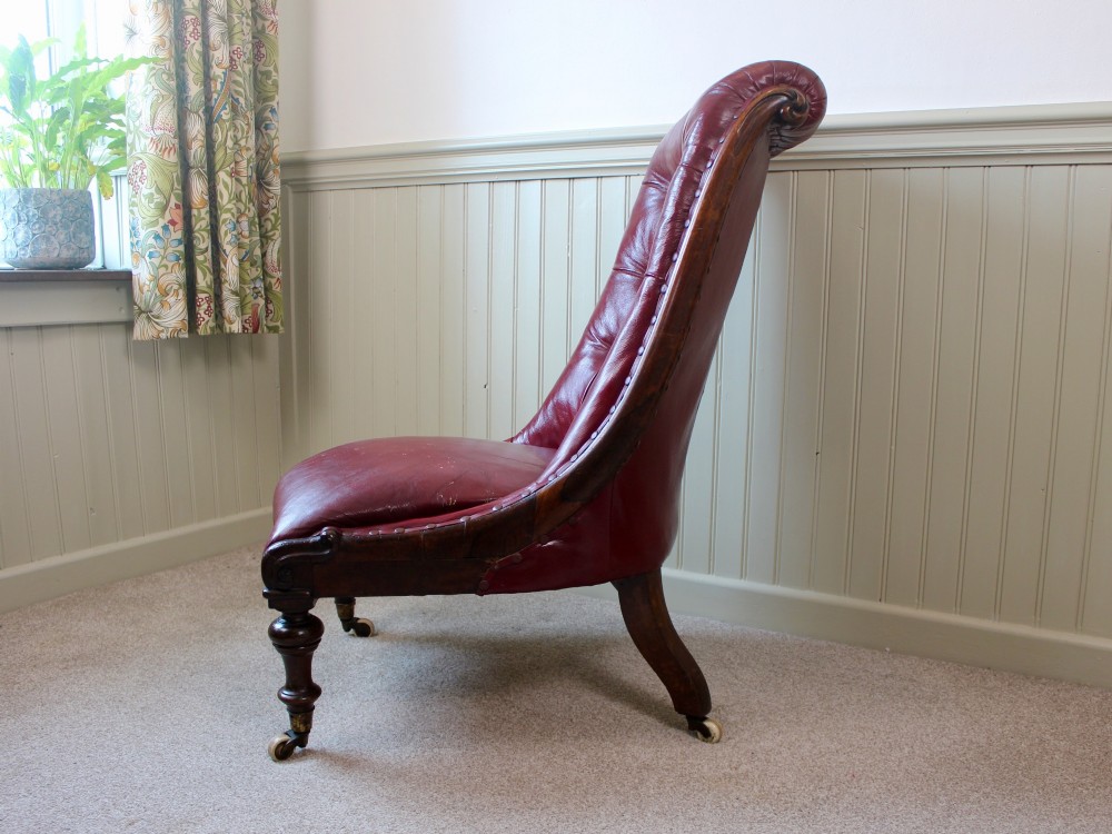 An early Victorian slipper chair featuring deep-buttoned red leather upholstery, a rosewood frame, and turned front legs on brass castors. View VI