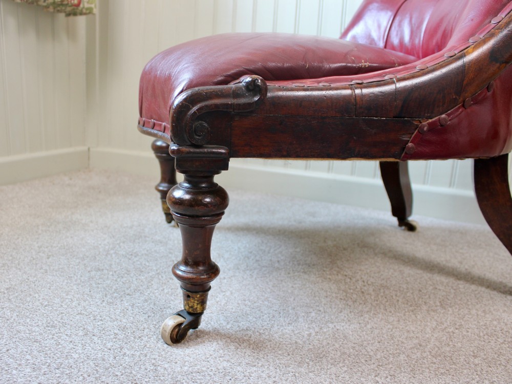 An early Victorian slipper chair featuring deep-buttoned red leather upholstery, a rosewood frame, and turned front legs on brass castors. View VII