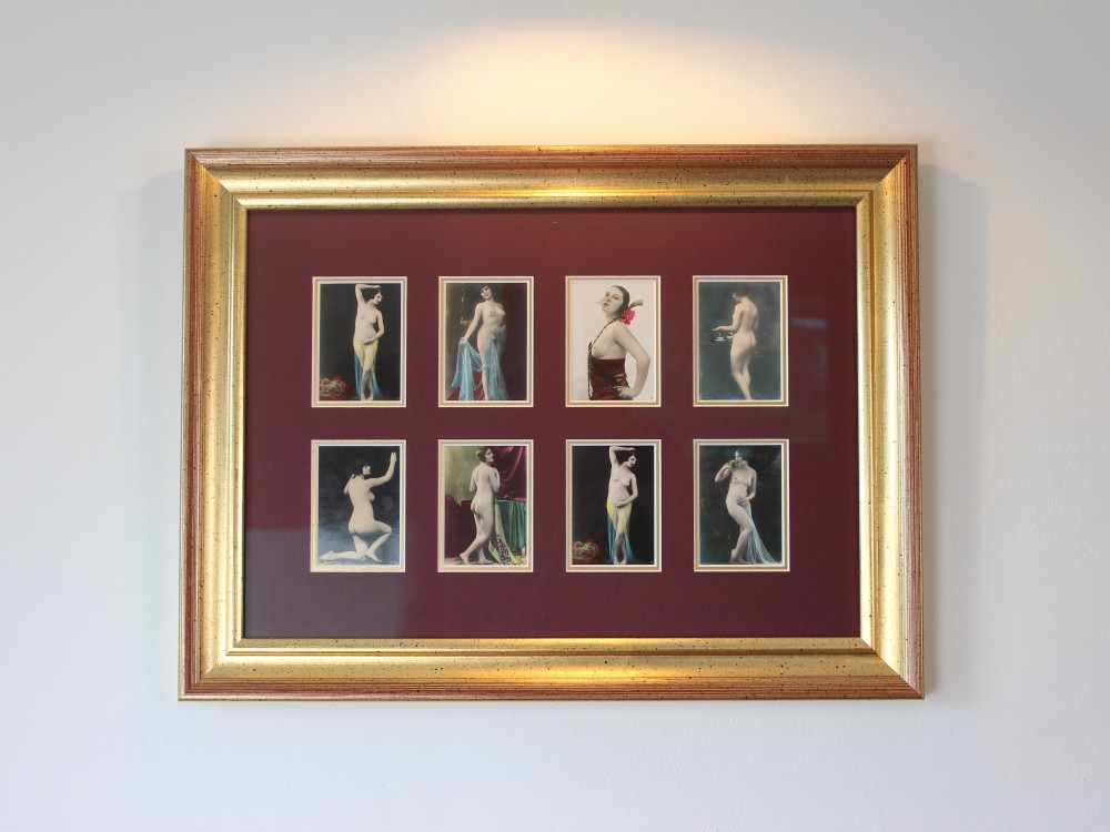 An array of eight 1920s hand-tinted female nude photographs mounted on a burgundy mat within a moulded gold frame.