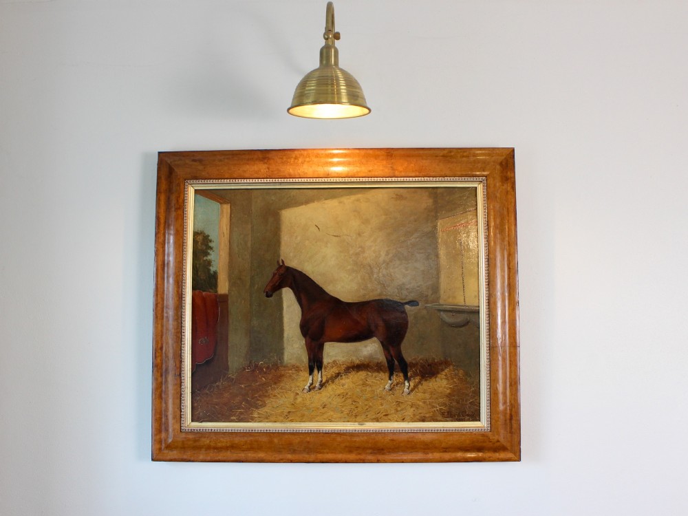 An antique oil painting depicting a bay horse standing inside a straw-filled stable within a wide timber frame.